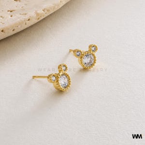 May include: A pair of gold-colored stud earrings in the shape of a mouse. Each earring features a large, clear, round gemstone in the center, surrounded by a decorative border. Two smaller gemstones form the ears. The earrings are on a neutral background.