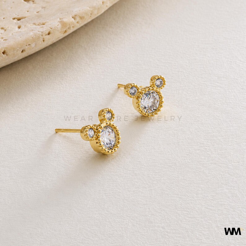 May include: A pair of gold-colored stud earrings in the shape of a mouse. Each earring features a large, clear, round gemstone in the center, surrounded by a decorative border. Two smaller gemstones form the ears. The earrings are on a neutral background.