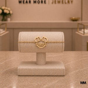 Pode incluir: Uma pulseira dourada com o nome "ISABELLA" &eacute; exibida em um expositor de joias bege. A pulseira apresenta um design inspirado no Mickey Mouse. O texto "WEAR MORE | JEWELRY" &eacute; vis&iacute;vel ao fundo.