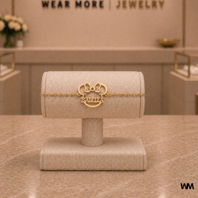 Pode incluir: Uma pulseira dourada com o nome "ISABELLA" &eacute; exibida em um expositor de joias bege. A pulseira apresenta um design inspirado no Mickey Mouse. O texto "WEAR MORE | JEWELRY" &eacute; vis&iacute;vel ao fundo.