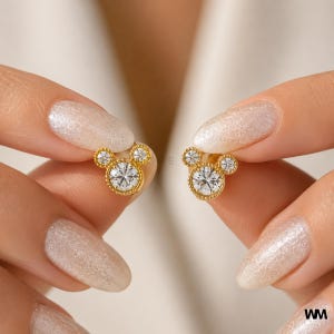 May include: A pair of gold-colored stud earrings, each featuring a large, round, clear gemstone center, surrounded by smaller clear gemstones, forming a Mickey Mouse head shape. The earrings are held between fingers against a soft, neutral background.