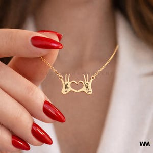May include: Gold-colored necklace with a pendant shaped like two hands forming a heart. The names "John" and "Sarah" are engraved on the pendant. The necklace is on a delicate chain.