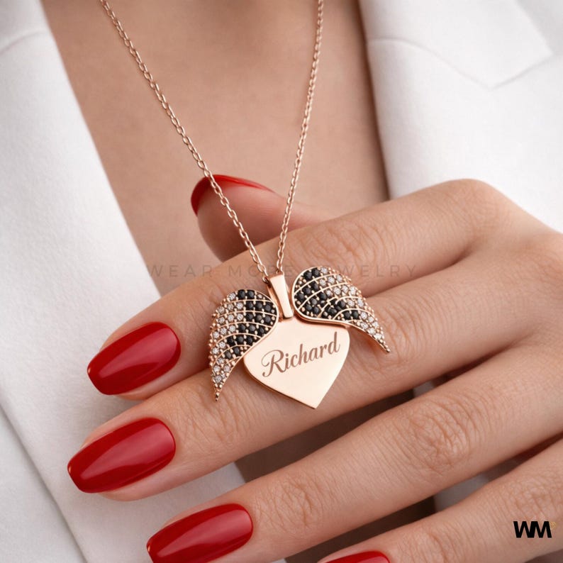 May include: A rose gold necklace with a heart-shaped pendant and angel wings. The heart is engraved with the name "Richard". The wings are embellished with black and clear crystals. The necklace is displayed on a person's hand with red nail polish.