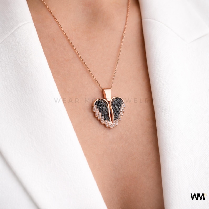 May include: A rose gold necklace with a heart-shaped pendant. The pendant features black and white stones arranged in a wing design. The necklace is worn against a white blazer.