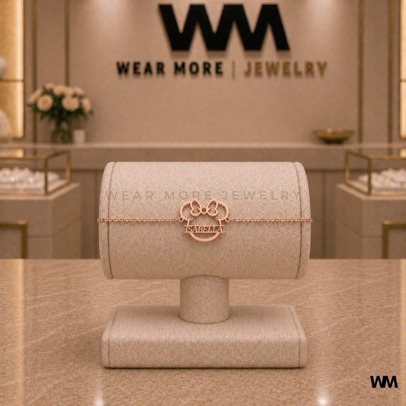 Pode incluir: Pulseira em ouro rosa com um design inspirado na Minnie Mouse. A pulseira apresenta um berloque com o nome "ISABELLA" inscrito dentro de uma moldura circular. O texto "WEAR MORE | JEWELRY" &eacute; vis&iacute;vel no fundo.
