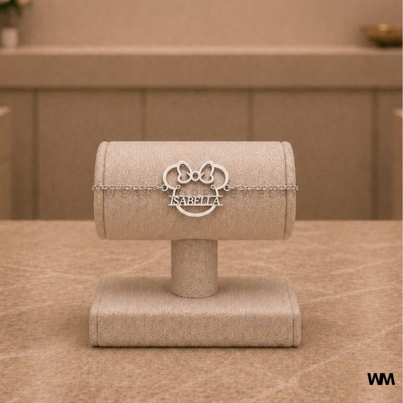 Pode incluir: Uma pulseira prateada com um design inspirado na Minnie Mouse, apresentando um la&ccedil;o e o nome "ISABELLA" em uma moldura circular. A pulseira &eacute; exibida em um suporte de joias bege, mostrando sua corrente delicada e detalhes intrincados.