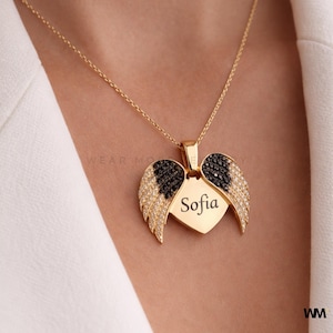 May include: Gold-colored necklace with a heart-shaped pendant and angel wings. The wings are adorned with black and clear crystals. The pendant has the name "Sofia" engraved on it. The necklace is worn on a white top.