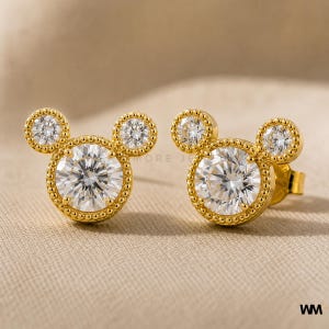 May include: A pair of gold-colored stud earrings in a Mickey Mouse design. Each earring features a large, round, clear gemstone surrounded by a beaded border, with two smaller gemstones above. The earrings are set against a neutral background.