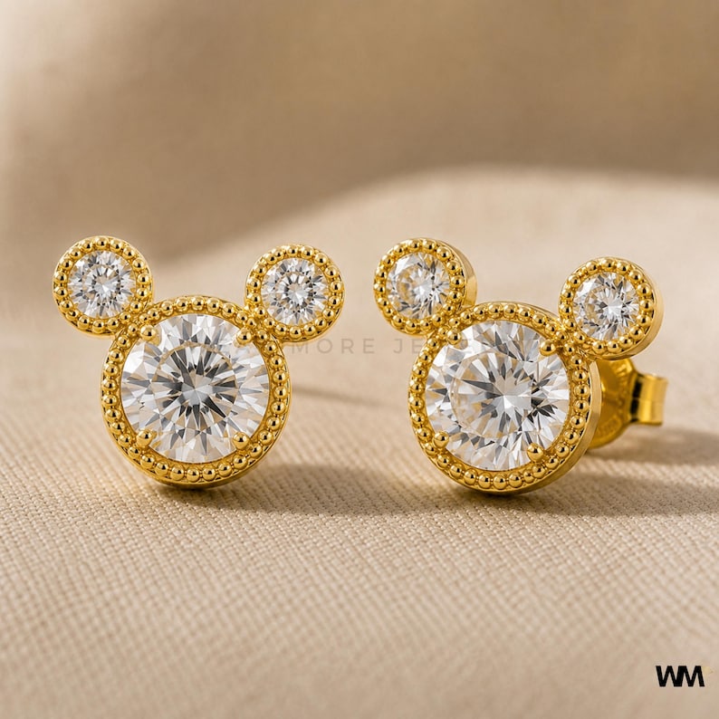 May include: A pair of gold-colored stud earrings in a Mickey Mouse design. Each earring features a large, round, clear gemstone surrounded by a beaded border, with two smaller gemstones above. The earrings are set against a neutral background.