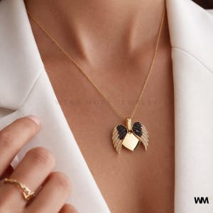 May include: A gold-colored necklace with a pendant shaped like a heart with angel wings. The wings are adorned with black and clear gemstones. The necklace is worn on a person wearing a white blazer. The text "WEAR MORE JEWELRY" is visible.