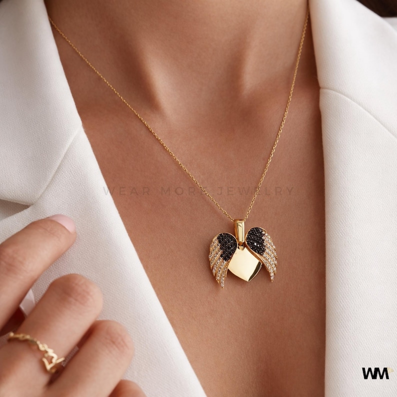 May include: A gold-colored necklace with a pendant shaped like a heart with angel wings. The wings are adorned with black and clear gemstones. The necklace is worn on a person wearing a white blazer. The text "WEAR MORE JEWELRY" is visible.