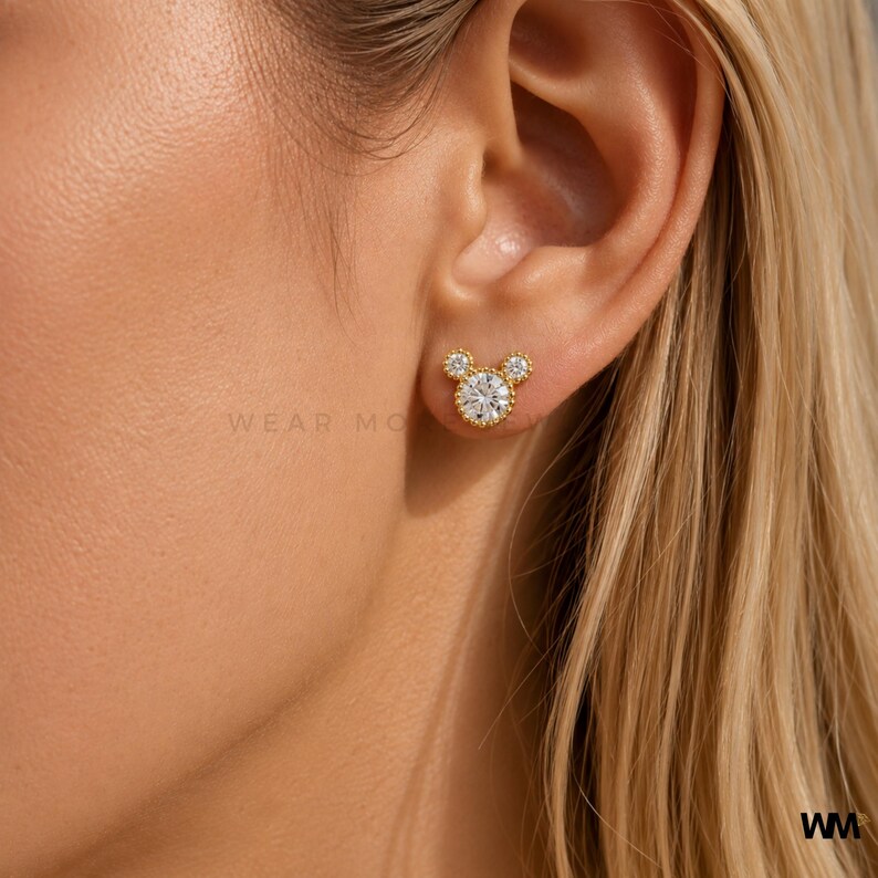 May include: Gold-colored stud earrings in a Mickey Mouse design, featuring a large, round, clear stone center with smaller stones on each side. The earrings are set against a person's ear, with the text "WEAR MO" visible.