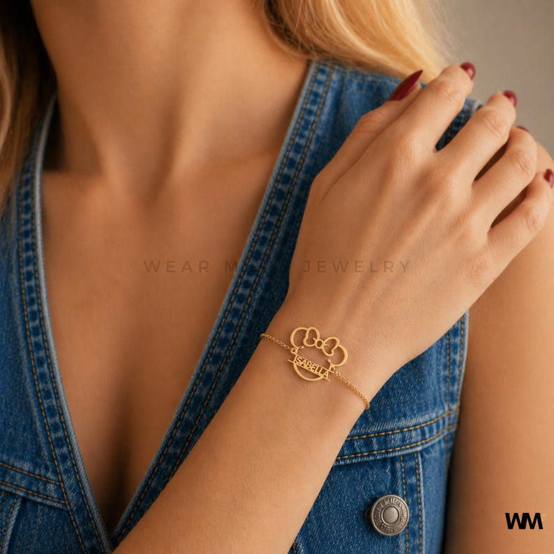 Pode incluir: Pulseira dourada com um pendente em forma de la&ccedil;o e o nome "Isabella". A pulseira est&aacute; presa a uma corrente fina. A imagem tamb&eacute;m mostra o texto "WEAR ME JEWELRY" e as iniciais "WM".