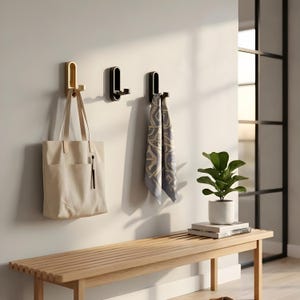 Op de afbeelding: Drie wandhaken in goud en zwart, met een beige draagtas en een sjaal met patroon. Een houten bank met een plant in een pot en boeken staat eronder. De scène speelt zich af tegen een neutrale muur.