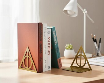 Könnte beinhalten: Ein Paar goldfarbene Buchstützen aus Metall, jede mit einem dreieckigen Rahmen, der einen Kreis und eine vertikale Linie umschließt. Eine Buchstütze stützt drei Bücher, während die andere leer steht und die geometrische Metallkonstruktion zeigt.