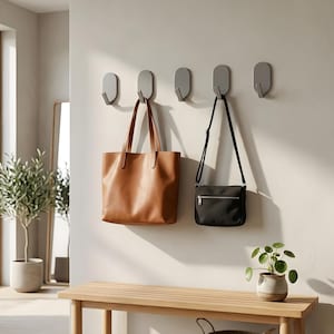 Op de afbeelding: Een wandoplossing met vijf grijze haken. Een bruine leren tote bag en een zwarte crossbodytas hangen aan de haken. Een houten bank en een potplant maken de inrichting compleet.