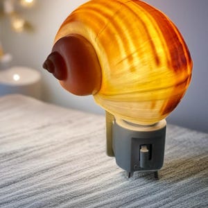 Puede incluir: Una luz de noche con forma de concha con un interior cálido y brillante. La concha es una mezcla de tonos amarillos y naranjas, con un acento marrón. La luz de noche tiene una base gris con un interruptor de encendido/apagado. Una pieza de iluminación decorativa única.