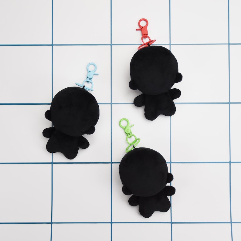 May include: Three black plush keychains shaped like a figure with a round head and small arms and legs. Each keychain has a different colored clip: red, blue, and green. The keychains are on a white tile background with blue lines.