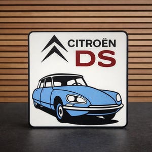 May include: Square sign featuring a light blue Citroën DS car illustration. The sign includes the Citroën logo and the text "CITROËN DS" in black and maroon. The car is set against a white background with a black border.