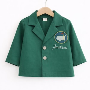 May include: A green blazer with a classic collar and two silver buttons. The blazer features an embroidered patch with a map of the United States and the name "Jackson". The blazer is hanging on a wooden hanger.