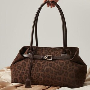 May include: A brown handbag with a leopard print design. The bag has two tall handles and a strap with a gold-colored buckle. The bag is made of a soft material and has a small, dangling leather piece.