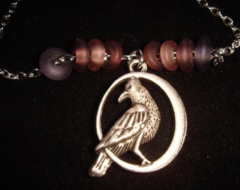May include: Silver-toned raven pendant necklace with a detailed raven design within a circular frame. The necklace features a chain and is adorned with purple and pink glass beads. The background is black.