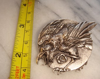 May include: Silver-toned metal pin featuring a detailed, three-dimensional design of a raven with its wings spread. The raven is perched on a circular base.