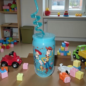 Pu&ograve; includere: Un bicchiere di plastica azzurro con coperchio e cannuccia riccia. Il bicchiere presenta personaggi del film Toy Story. Vari blocchi di giocattoli e un'auto giocattolo rossa sono sparsi intorno al bicchiere.