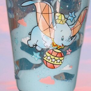 Puede incluir: Un vaso transparente con líquido azul y un diseño del elefante Dumbo. El diseño incluye estrellas, nubes y una bola colorida. El vaso está lleno de purpurina y tiene una estética caprichosa y de dibujos animados.