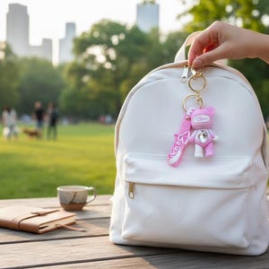 May include: A white backpack with a gold zipper, held on a wooden table. A pink bear keychain with a rhinestone is attached to the zipper. A notebook and a cup of coffee are also on the table. The background shows a park with people.