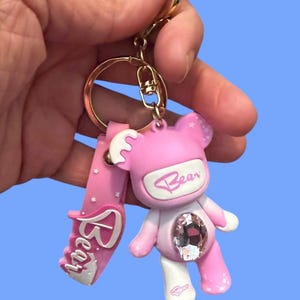 May include: Pink bear keychain with a large crystal embedded in the center. The bear has white accents and the word "Bear" is written on the front. The keychain has a gold-colored ring and a pink strap with the word "Bear" on it.
