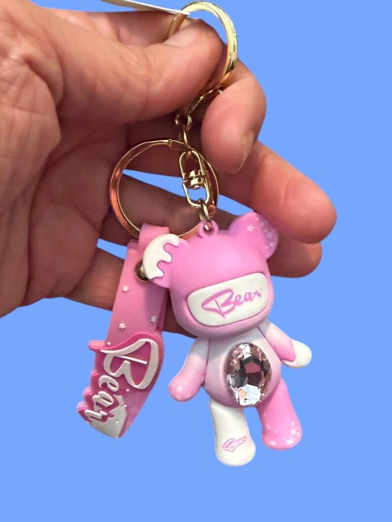 May include: Pink bear keychain with a large crystal embedded in the center. The bear has white accents and the word "Bear" is written on the front. The keychain has a gold-colored ring and a pink strap with the word "Bear" on it.