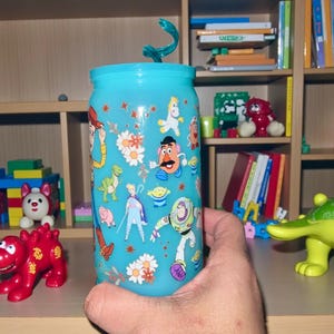 Pu&ograve; includere: Un bicchiere azzurro con coperchio abbinato e cannuccia a spirale. Il bicchiere &egrave; decorato con personaggi dei cartoni animati e motivi floreali. Il bicchiere &egrave; tenuto davanti a una mensola di legno con giocattoli.