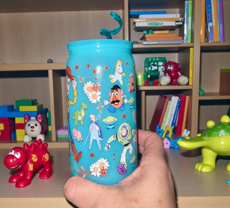 Pu&ograve; includere: Un bicchiere azzurro con coperchio abbinato e cannuccia a spirale. Il bicchiere &egrave; decorato con personaggi dei cartoni animati e motivi floreali. Il bicchiere &egrave; tenuto davanti a una mensola di legno con giocattoli.