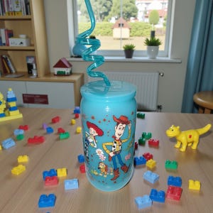 Pu&ograve; includere: Un bicchiere azzurro con coperchio e cannuccia a spirale. Il bicchiere presenta i personaggi del film Toy Story, tra cui Woody e Jessie. Blocchi colorati e una statuina di dinosauro giallo sono sparsi.