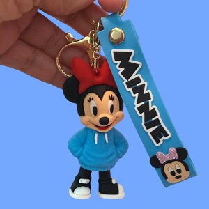 May include: A Minnie Mouse keychain featuring a blue hoodie, red bow, and black shoes. The keychain includes a blue strap with the name "MINNIE" in white letters and a Minnie Mouse face.