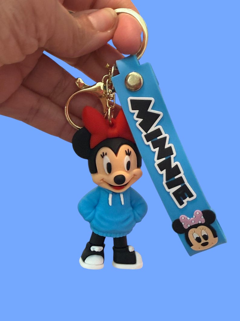 May include: A Minnie Mouse keychain featuring a blue hoodie, red bow, and black shoes. The keychain includes a blue strap with the name "MINNIE" in white letters and a Minnie Mouse face.