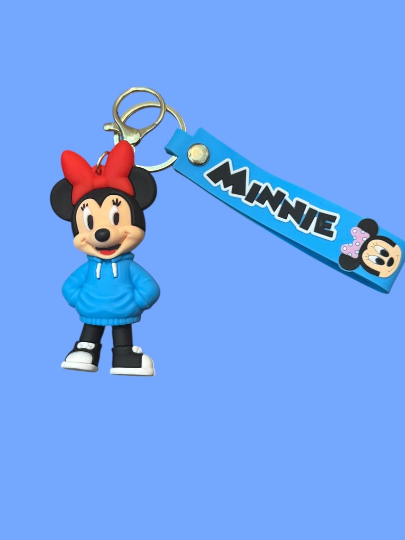 May include: A Minnie Mouse keychain featuring a cartoon figure wearing a blue hoodie and red bow. The keychain includes a blue strap with the name "Minnie" in white letters and a gold-colored clasp.