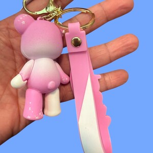 May include: A pink bear keychain with a gold-colored clasp and ring. The bear has white accents and star details. A pink strap with a white edge is attached to the ring.