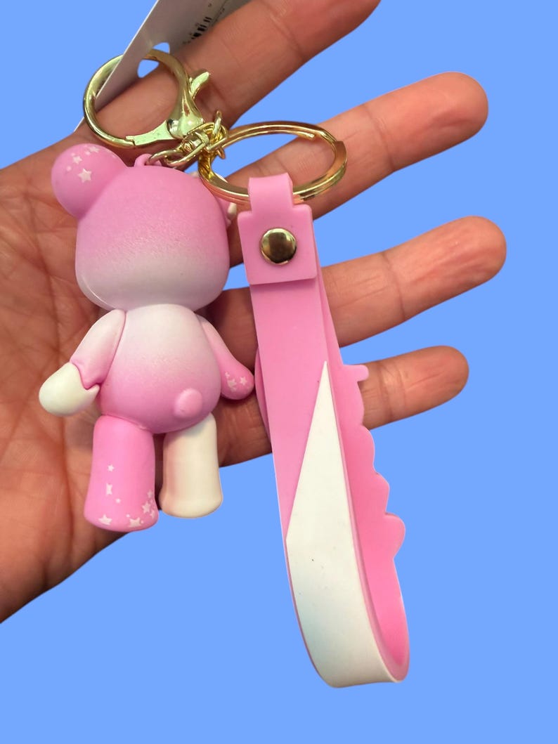 May include: A pink bear keychain with a gold-colored clasp and ring. The bear has white accents and star details. A pink strap with a white edge is attached to the ring.