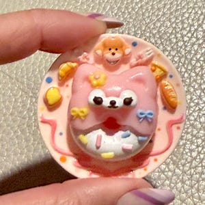 May include: A pink, circular charm featuring a cartoon cat design with a donut-shaped mouth. The charm has a light pink background with a small monkey figure, yellow flowers, and blue bows. The charm is held by a person.