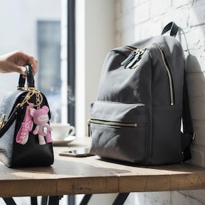 May include: A black handbag with a long strap and gold hardware is held by a person. A pink bear keychain hangs from the handle. A gray backpack with gold zippers sits on a wooden table.