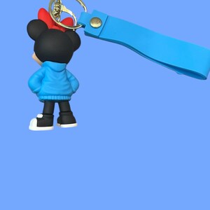 May include: A Minnie Mouse keychain featuring a black figure with a red bow, blue jacket, and black shoes with white soles. The keychain has a gold ring and a light blue strap.