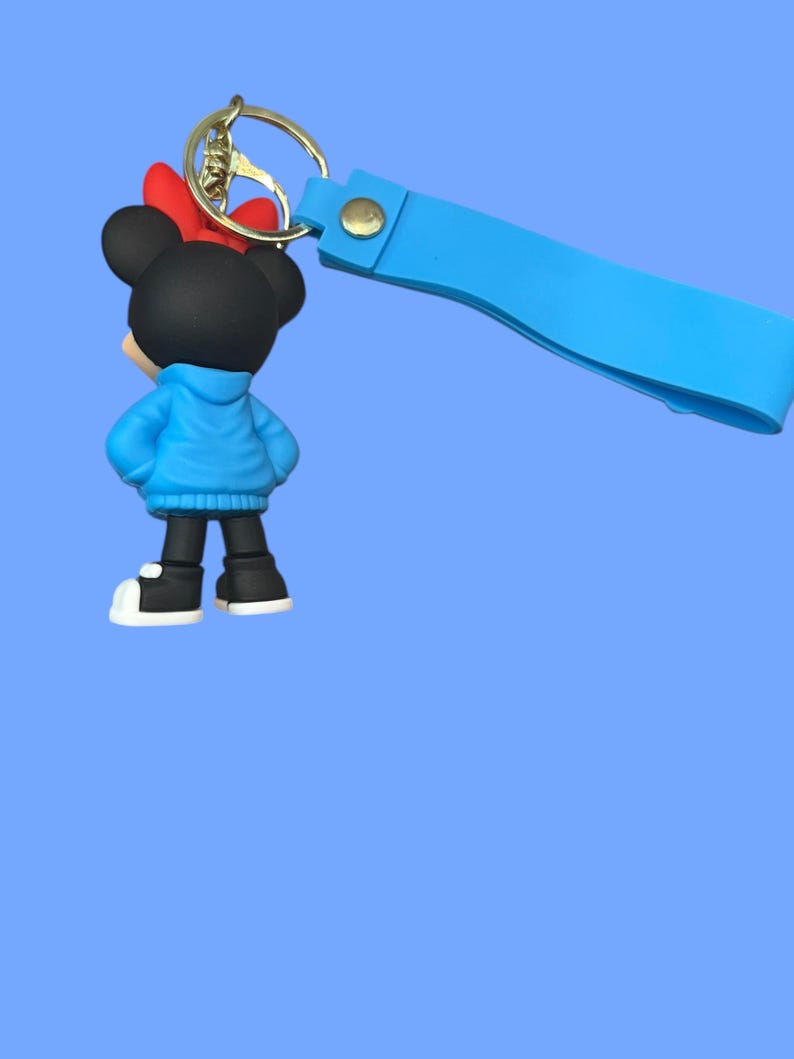 May include: A Minnie Mouse keychain featuring a black figure with a red bow, blue jacket, and black shoes with white soles. The keychain has a gold ring and a light blue strap.