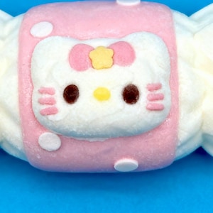May include: A pink and white candy shaped like a wrapped treat, featuring a Hello Kitty design. The candy has a pink center with a white Hello Kitty face, complete with a pink bow and yellow star. The candy is on a blue background.