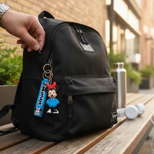 May include: Black backpack with a Minnie Mouse keychain. The keychain has a blue strap with the word "MINNIE" and a Minnie Mouse figure wearing a blue hoodie. A person's hand is opening the zipper. Headphones and a water bottle are on a wooden bench.