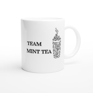 Peut inclure: Un mug en céramique blanche avec le texte imprimé en noir TEAM MINT TEA sur le côté gauche et une illustration graphique noire représentant un verre rempli de feuilles de menthe avec de la vapeur s'élevant sur le côté droit du mug.