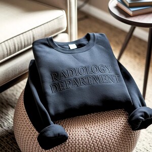 Könnte beinhalten: Ein marineblaues Sweatshirt mit dem gestickten Schriftzug "RADIOLOGY DEPARTMENT" auf der Brust. Das Sweatshirt liegt gefaltet auf einem gewebten Hocker. Die Ärmel sind umgeschlagen.