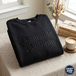May include: Black crew neck sweatshirt featuring the embroidered phrase "Probably Nothing" in a gothic font. The sweatshirt is folded on a neutral surface. A coffee cup and books are in the background.