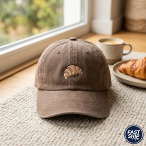 Könnte beinhalten: Braune Baseballkappe mit gebogenem Schirm und kleinem, gesticktem Croissant-Design. Die Kappe besteht aus einem weichen, strukturierten Stoff und hat ein klassisches Sechs-Panel-Design. Die Kappe ist neutral gefärbt.